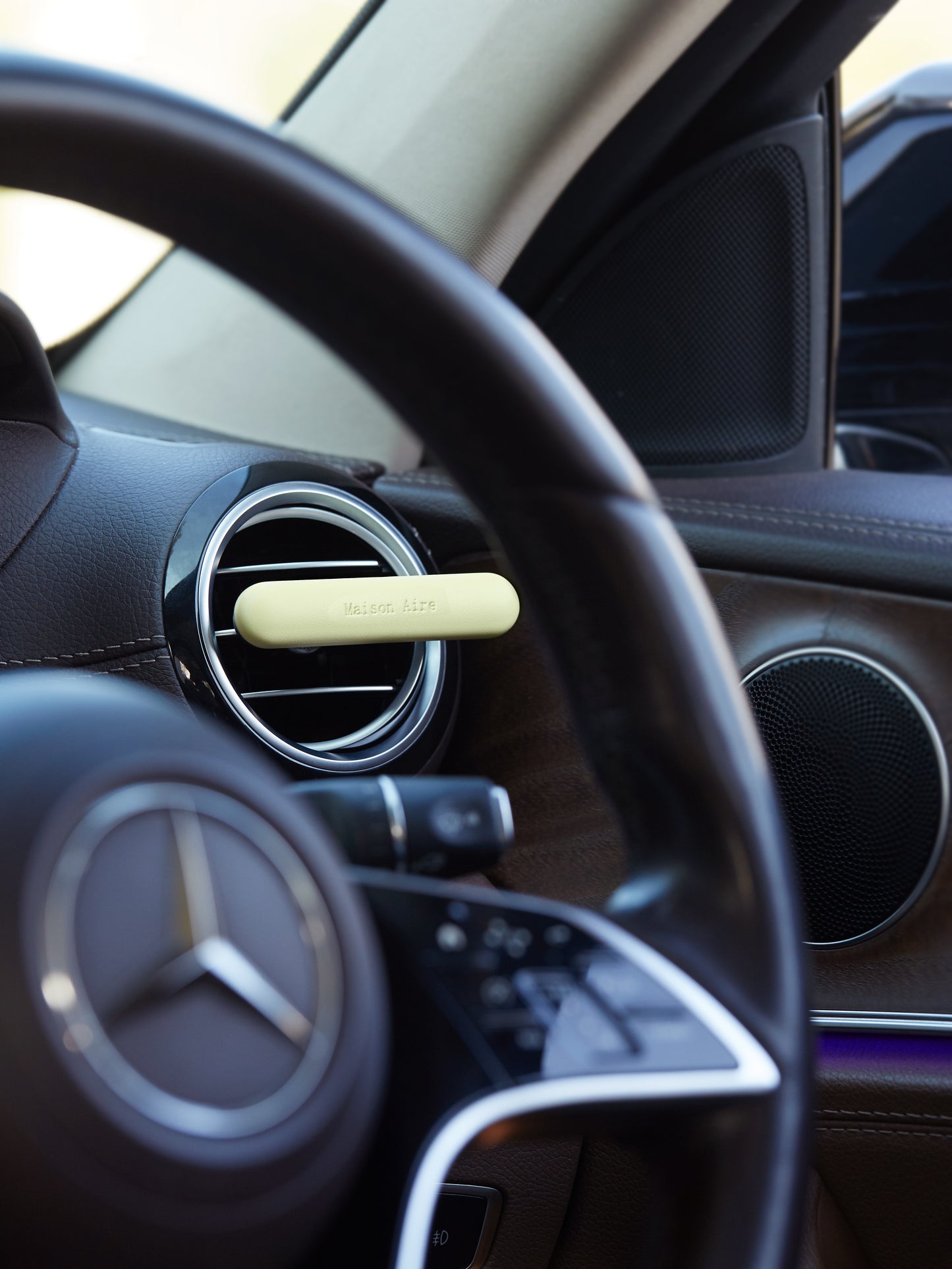 Car interior with luxury brown leather dashboard, Mercedes steering wheel, and a butter yellow leather Maison Aire air freshener diffuser on air vent
