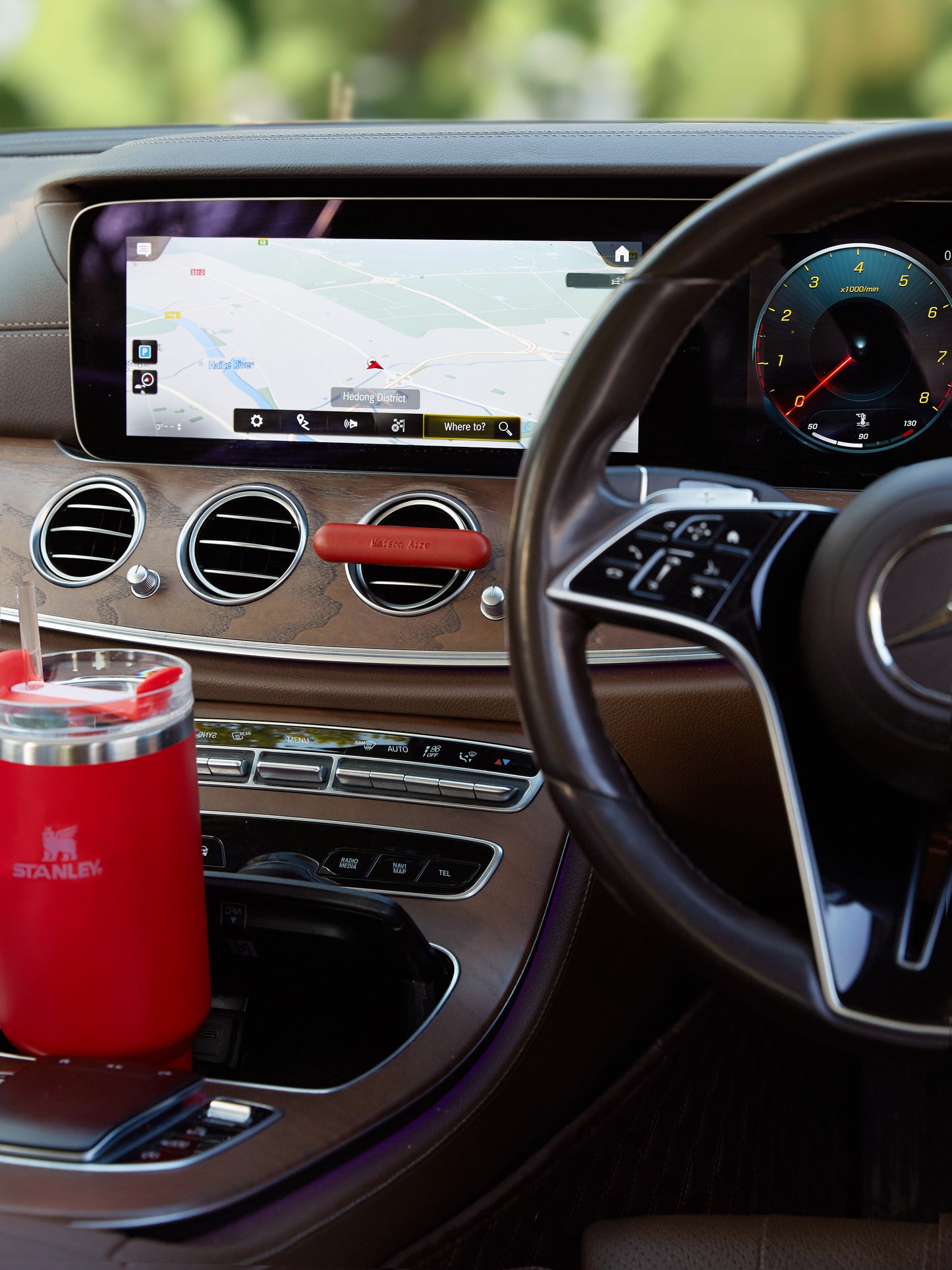 Mercedes-Benz Luxury car interior dashboard with navigation screen, steering wheel, and red Stanley mug in cup holder and red leather Maison Aire car diffuser 