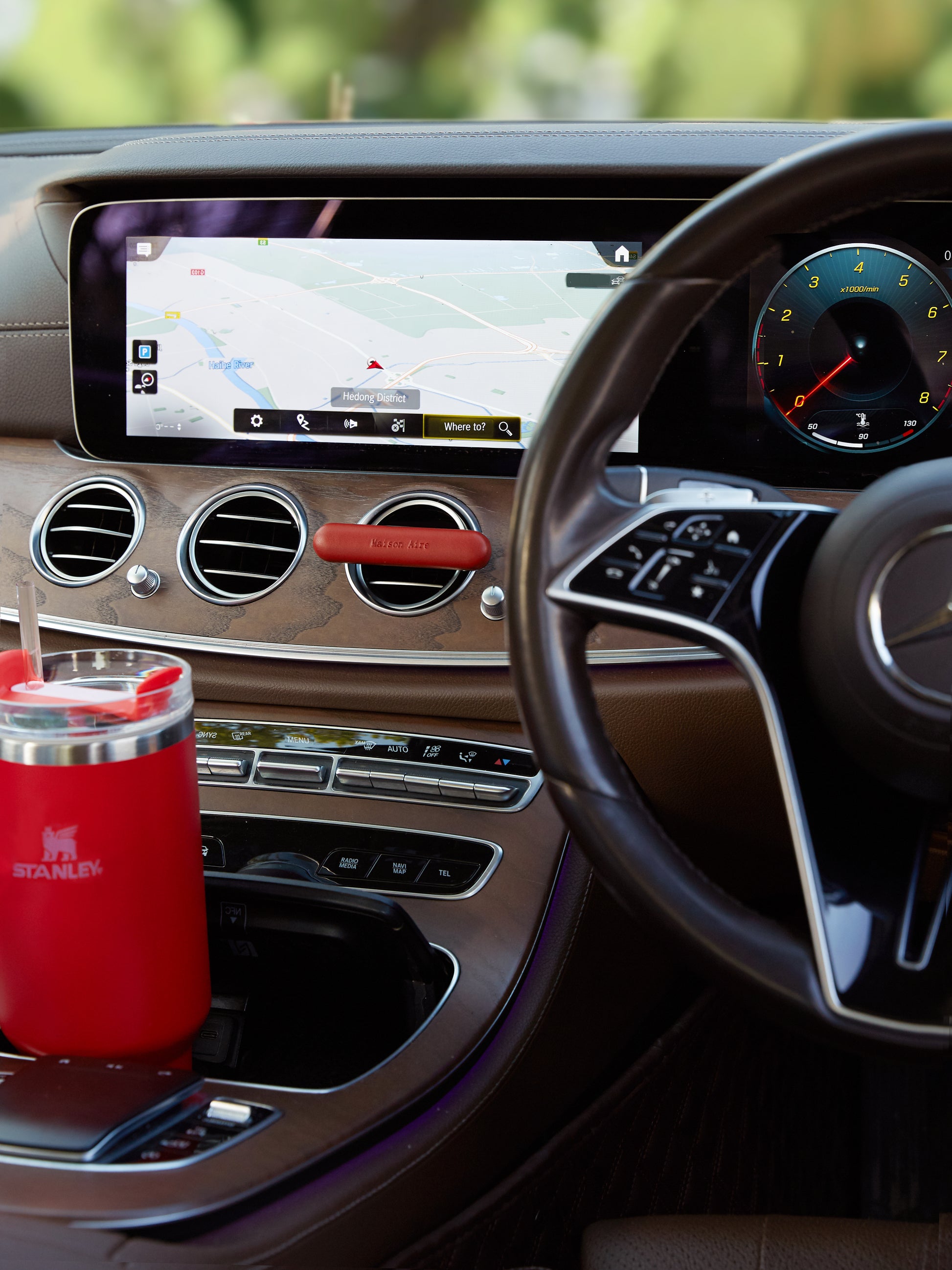 Mercedes-Benz Luxury car interior dashboard with navigation screen, steering wheel, and red Stanley mug in cup holder and red leather Maison Aire car diffuser 