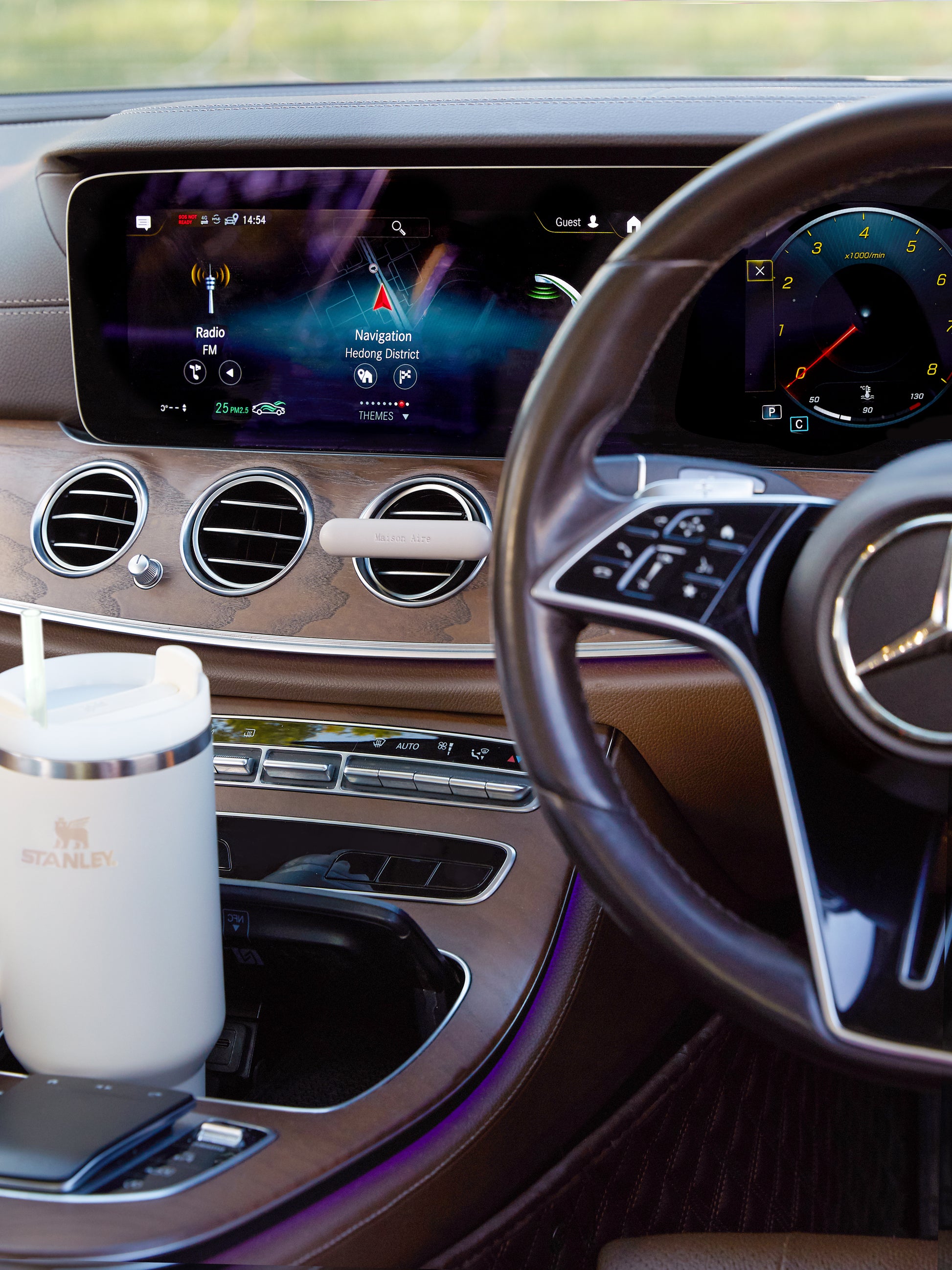 Mercedes-Benz Luxury car interior dashboard with navigation screen, steering wheel, and grey Stanley mug in cup holder and a grey leather Maison Aire car diffuser 
