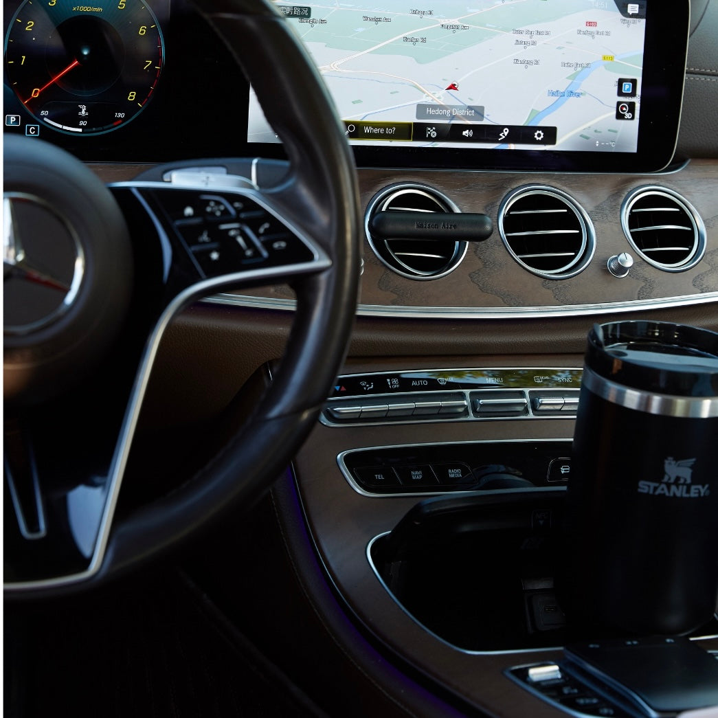 Luxury car dashboard with steering wheel, digital speedometer, navigation screen, air vents, and Stanley coffee mug